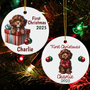 May include: Two white ceramic Christmas ornaments with gold hanging strings. One features a brown puppy in a gift box, the text "First Christmas 2025" and "Charlie". The other shows a puppy with a red bow, the text "First Christmas", "Charlie" and "2025".
