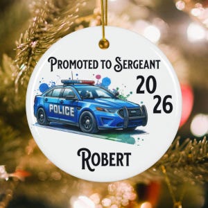 May include: A white ceramic ornament with a gold string features a blue police car illustration. Text reads "Promoted to Sergeant 2026" above the car, with "Robert" below. The ornament is round and has a gold-colored top.