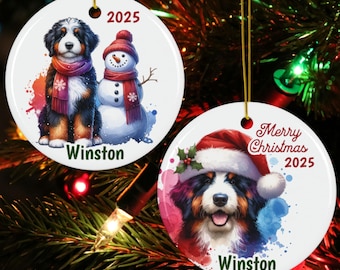 May include: Two round white ceramic ornaments featuring Bernese Mountain Dog illustrations. One shows a dog with a snowman, the year 2025, and the name Winston. The other shows a dog in a Santa hat, the text Merry Christmas 2025, and the name Winston.