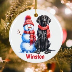 May include: A round white ceramic ornament featuring a snowman wearing a red patterned hat and scarf next to a black dog wearing a matching red patterned scarf. The name Winston is printed in red letters at the bottom of the ornament.