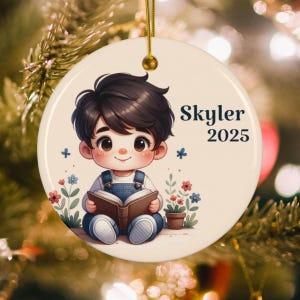 May include: Round ceramic ornament featuring a cartoon illustration of a child reading a book, with the name "Skyler" and the year "2025" printed in black. The ornament has a gold string for hanging and is decorated with flowers.