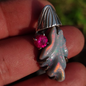 Può includere: Ciondolo in argento con design a cappello di fungo, una gemma rosa e una pietra simile all'opale. Il ciondolo ha una forma organica e colori iridescenti.