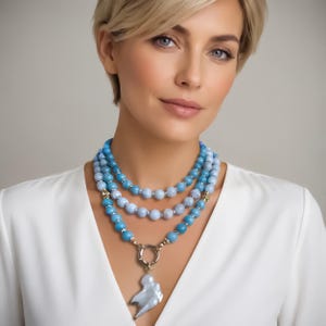 Puede incluir: Un collar de tres hilos con cuentas azul claro y turquesa. El collar tiene un cierre dorado y un colgante nacarado. El collar se lleva sobre una blusa blanca.