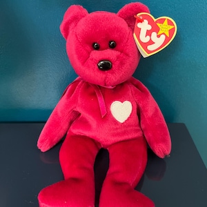 May include: Pink Beanie Baby teddy bear with a white heart on its chest. The bear has black eyes and a black nose. The tag reads "Beanie Babies" and "Ty".