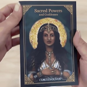 A person's hands holding a book titled "Sacred Powers and Goddesses" by Claudia Oliveros.