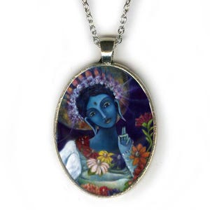 May include: An oval silver-toned pendant necklace featuring a colorful artistic illustration of a blue-skinned figure with a floral crown and a third eye, surrounded by vibrant flowers against a dark, circular background.