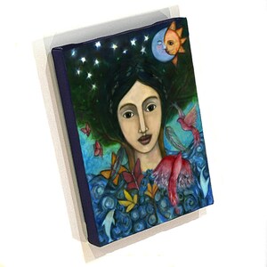 May include: A painting of a woman with dark hair and brown eyes. She is surrounded by a blue and green background with stars, birds, and leaves. The woman is wearing a blue dress with a pink bird on her shoulder.