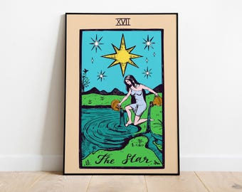 May include: A framed print of The Star tarot card. The artwork depicts a woman pouring water from two golden jugs into a river. A large yellow star and smaller stars are in the blue sky above. The card is framed in black.