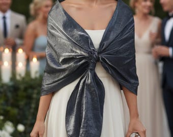 May include: A woman in a white dress with a dark grey, shimmering shawl tied in a bow. She holds a small, silver clutch. The background shows other people, suggesting a formal occasion. The shawl adds a touch of elegance.