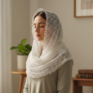 Op de afbeelding: Een witte kanten hoofdbedekking met een delicaat bloemenpatroon en een decoratieve rand van kleine, ronde pompons langs de zomen. De stof is halfdoorzichtig en valt soepel over het hoofd en de schouders voor een zachte, gelaagde uitstraling.