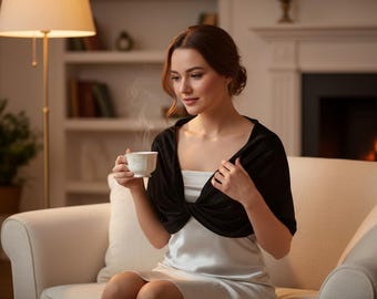 May include: A woman seated on a cream-colored sofa, wearing a black velvet wrap and a white dress, holds a white teacup with steam rising. A lit fireplace and a lamp are in the background.