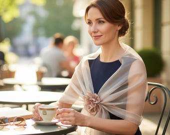 May include: A woman wearing a navy blue dress and a sheer, light beige shawl with a large flower accent. She is seated at a cafe table, holding a teacup. Sunglasses and a book rest on the table.