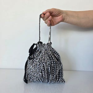 May include: A black beaded drawstring bag with a black ribbon tie. The bag is covered in small, shiny beads in shades of silver and white. The handle is made of a chain of small beads.