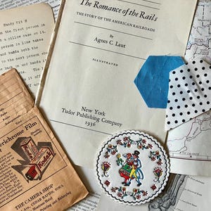 K&ouml;nnte beinhalten: Ein Vintage-Buch mit dem Titel "The Romance of the Rails: The Story of the American Railroads" von Agnes C. Laut, illustriert und ver&ouml;ffentlicht 1936 von Tudor Publishing Company in New York. Der Buchumschlag ist wei&szlig; mit schwarzem Text. Ein blauer und wei&szlig;er Stoffrest mit wei&szlig;en Punkten befindet sich auf dem Umschlag.