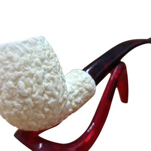 Peut inclure: Une pipe à tabac avec un foyer texturé blanc cassé et un tuyau marron foncé. La pipe repose sur un support rouge brillant, créant un contraste saisissant. Le foyer présente un motif irrégulier unique.