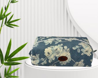 May include: A rectangular, floral-patterned cosmetic bag in shades of blue and white, with a small brown label. The bag is displayed on a white pedestal, with bamboo stalks and leaves in the background.