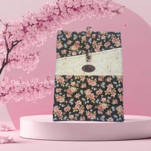 May include: A floral pattern book cover with a black background and pink and white flowers. The cover has a cream-colored horizontal stripe with a brown leather tag that has the initials "Bg". The cover is standing on a pink pedestal.