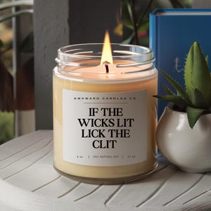 May include: A clear glass candle jar with a white label that reads "AWKWARD CANDLES CO. IF THE WICKS LIT LICK THE CLIT 8 oz | 100% NATURAL SOY | 50 HR"
