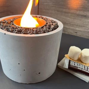 May include: A cylindrical gray concrete fire pit with a central metal burner containing a bright orange flame surrounded by dark volcanic rocks. Next to the fire pit are two white marshmallows on a graham cracker and a Hershey's milk chocolate bar.