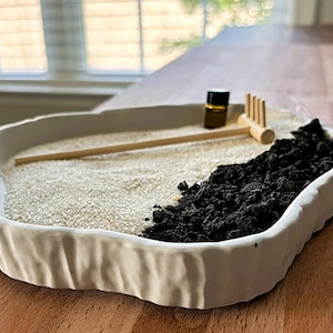 May include: A white, irregularly shaped ceramic tray containing white sand and a section of black volcanic rock. A small wooden rake and a tiny dark glass bottle are placed on the sand. The tray has a textured, uneven outer edge.