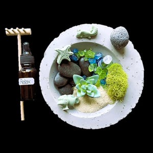 May include: A small concrete bowl with a sand base, green moss, small stones, a starfish, a succulent plant, and a small green figurine. The bowl is on a black background. The word "Relax" is written on a small bottle.