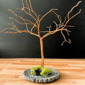 May include: A wire sculpture of a tree with a brown trunk and branches. The tree is sitting on a gray, circular base with green moss.