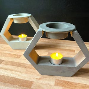 May include: Two geometric concrete candle holders. Each holder has a hexagonal shape with a circular indentation at the top for a candle and a lower section to hold a lit tea light. One is light gray, the other is off-white.