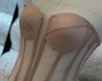 May include: A nude-colored corset with a structured bust and sheer panels. The corset features vertical boning and a delicate, feminine design. The garment is displayed on a mannequin, showcasing its shape and construction.