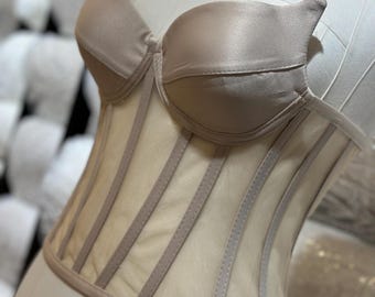 May include: A beige mesh corset with a satin top and a boned bodice. The corset has a fitted silhouette and a smooth, silky texture.