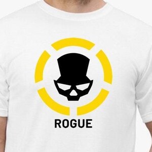 May include: White t-shirt with a black and yellow logo. The logo is a skull wearing a top hat inside a circle with three yellow rings. The text "ROGUE" is below the logo.