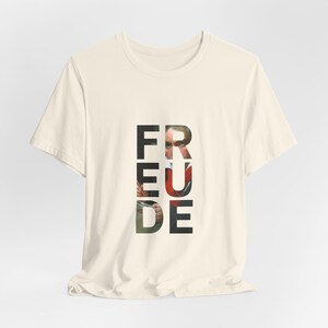 Könnte beinhalten: Cremefarbenes T-Shirt mit dem Wort "FREUDE" in vertikaler, fetter, schwarzer Schrift. Der Buchstabe "R" zeigt ein Teilbild eines Gesichts und roter Kleidung. Das Shirt ist aus weichem Material.