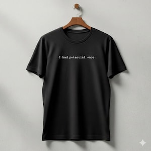 Può includere: T-shirt nero girocollo appeso a una gruccia di legno. La scritta bianca "I had potential once." è stampata al centro del petto. La maglietta è realizzata in un tessuto morbido.
