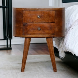 Könnte beinhalten: Ein Nachttisch aus Holz mit zwei Schubladen und einem abgerundeten Design. Der Nachttisch ist aus warmbraunem Holz gefertigt und hat einen modernen Stil aus der Mitte des Jahrhunderts. Er steht auf drei abgewinkelten Beinen und hat kleine, runde Schubladengriffe.