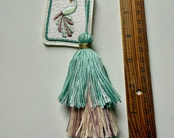 May include: Embroidered peacock keychain with a layered tassel. The keychain features a white tag with a peacock design in green, pink, and white embroidery. The tassel is in shades of green, tan, and pink, and is approximately 4 inches long.