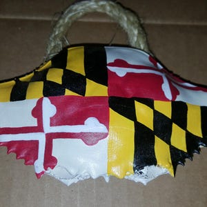 Puede incluir: Un adorno de caparazón de cangrejo pintado con la bandera del estado de Maryland. El caparazón es blanco con pintura roja, amarilla y negra. El adorno tiene un lazo de cuerda para colgar.