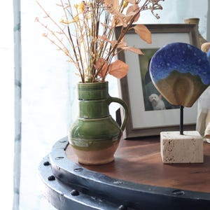 May include: A green ceramic vase with a handle, holding dried flowers and branches. The vase has a two-tone design, with a light brown base and a green upper section. The vase is on a dark wooden table.