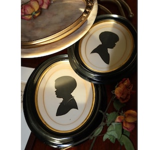 May include: Two oval framed silhouette portraits of a child in profile, set against a cream background with a gold inner border. The frames are black with a gold trim. Dried roses and a decorative tray are also visible.