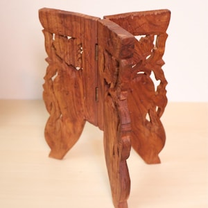 May include: A brown wooden folding stand featuring intricate carved floral patterns and cut-out details. The stand is constructed from two interlocking pieces connected by a metal hinge, forming a cross-shaped base designed to support objects.
