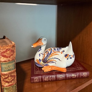 May include: A ceramic duck figurine with a white body, blue and orange floral patterns, and an orange beak. The duck is resting on a burgundy book with gold lettering. The figurine is displayed on a wooden shelf.