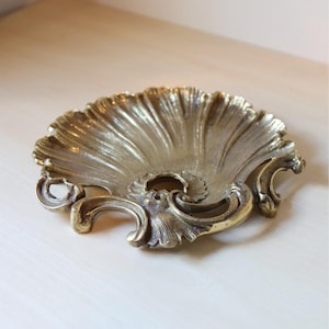 May include: Ornate, antique-style brass dish with a seashell design. The dish features a scalloped edge and intricate scrollwork. The brass has a warm, golden hue, and the dish is likely used for decorative purposes.