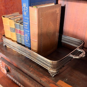 May include: A rectangular silver-toned metal tray with an ornate lattice-patterned gallery rail and decorative feet, holding a collection of five hardcover books, including a blue volume titled Grimm's Fairy Tales and a tan leather-bound book.