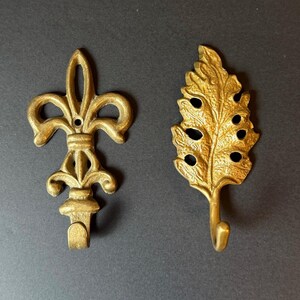 May include: Two gold-colored metal wall hooks. One hook features a symmetrical fleur-de-lis design with a central mounting hole. The second hook is shaped like a textured leaf with a curved hook extending from the bottom of the foliage.