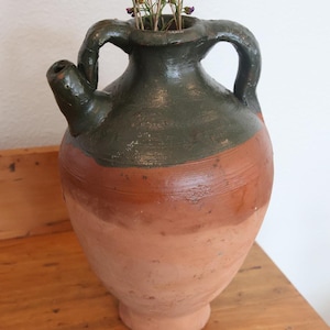 May include: A rustic, two-handled earthenware jug with a dark green top and a reddish-brown body. The jug has a spout and contains dried flowers. The jug is sitting on a wooden surface.