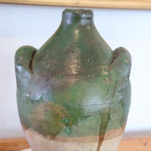 May include: Antique green ceramic jug with two side handles. The jug has a rounded body and a narrow neck. The glaze has a mottled appearance with shades of green and brown. The base is a lighter, unglazed color. A vintage decorative piece.