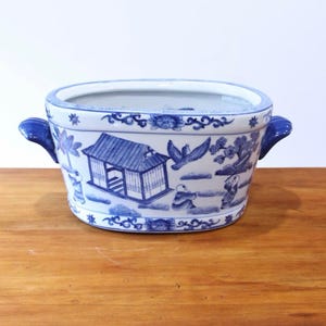 May include: An oval ceramic planter featuring a white base with blue hand-painted illustrations of a traditional house, a flying bird, and figures. The vessel includes two blue side handles and decorative floral borders along the top and bottom edges.