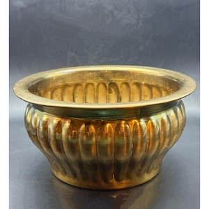May include: A round, antique brass planter with a fluted exterior. The planter has a wide, flat rim and a slightly curved base. The brass has a warm, golden hue and shows signs of age and use.