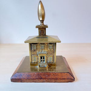 May include: A vintage brass miniature building with a spire, set on a wooden base. The building features detailed windows and a door. The brass has a warm, aged patina. The base is rectangular and made of dark wood.