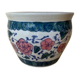 May include: A white ceramic bowl with a blue and green decorative border. The bowl features pink floral designs with green leaves. The rim of the bowl is white.