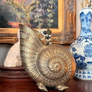 May include: A brass seashell-shaped vase with a textured surface, holding dried eucalyptus branches. The vase sits on a wooden surface, with a painting and a blue and white porcelain vase in the background.