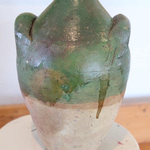 May include: A rustic, antique ceramic jug with a green glazed top and a tan base. The jug has two small handles and a rounded body. The glaze has a mottled appearance, with variations in color. The jug is likely used for decorative purposes.